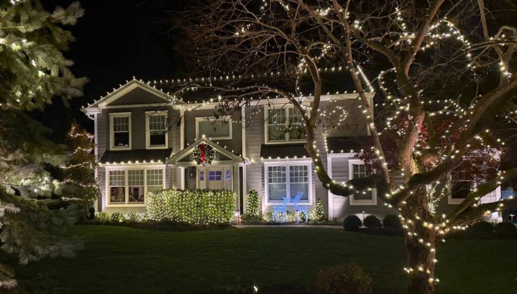 Two-story home at night with warm white roofline lights, a lit hedge, light-wrapped trees and a wreath over the front door